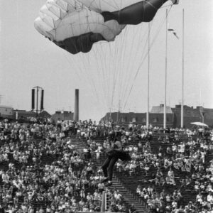 młody mężczyzna w kombinezonie z paralotnią na stadionie, w tle trybuny i drzewa