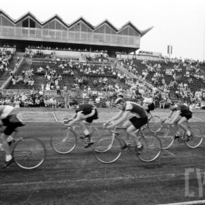 grupa mężczyzn w strojach sportowych ścigająca się na rowerach na tle kibiców siedzących na trybunach stadionu