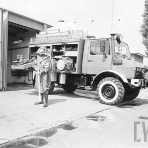 mężczyzna w kombinezonie strażackim; z tyłu strażacki wóz bojowy