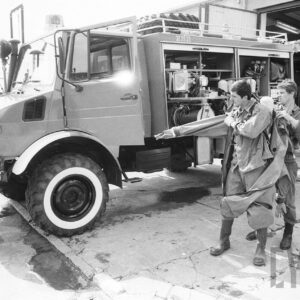 dwóch mężczyzn ubierających kombinezony strażackie; z tyłu strażacki wóz bojowy