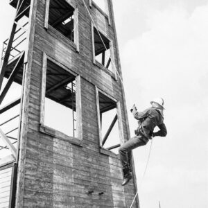 mężczyzna w kombinezonie i hełmie strażackim wspinający się po drabinie na drewnianą ścianę do ćwiczeń