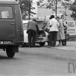 grupa ludzi stojąca przy samochodzie z otwartym bagażnikiem, w tle budynek mieszkalny z wielkiej płyty