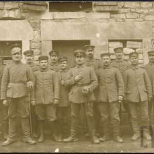 grupa mężczyzn w mundurach wojskowych, dwóch mężczyzn w pierwszym rzędzie podpiera się szablami, stoją na tle kamiennej ściany budynku z oknami