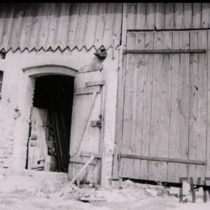 z lewej fragment ceglanego, murowanego budynku z otwartymi drewnianymi drzwiami, na lewo od nich małe okno, nad drzwiami pas przybitych pionowo desek, z prawej fragment drewnianego budynku