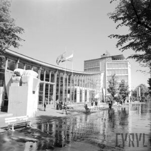 przeszklony budynek z dachem w kształcie delikatnej paraboli, spacerujący ludzie, część siedzi na ławkach przed budynkiem, z lewej pomnik w kształcie dwóch zaciśniętych pięści