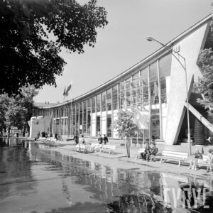 przeszklony budynek z dachem w kształcie delikatnej paraboli, spacerujący ludzie, część siedzi na ławkach przed budynkiem, w głębi pomnik w kształcie dwóch zaciśniętych pięści