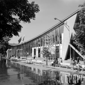 przeszklony budynek z dachem w kształcie delikatnej paraboli, spacerujący ludzie, część siedzi na ławkach przed budynkiem, w głębi pomnik w kształcie dwóch zaciśniętych pięści