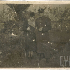 mężczyzna w mundurze podoficera stoi z kobietą ubraną w płaszcz i kapelusz na tle drzew i drewnianego płotu