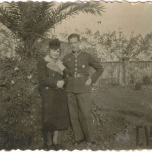 mężczyzna w mundurze podoficera stoi z kobietą ubraną w płaszcz i kapelusz na tle drzew i drewnianego płotu