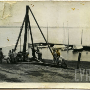 grupa żołnierzy w mundurach polowych przy drewnianej konstrukcji na tle jeziora i żaglówek 