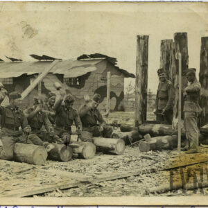 grupa mężczyzn w mundurach polowych na tle baraków wojskowych; czterech z nich siedzi przy belach drewna i uderza młotami (?), dwóch kolejnych stoi i przygląda się pozostałym
