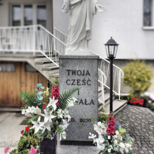 na kamiennym postumencie biała figura Chrystusa z rozłożonym dłońmi, na postumencie napis „Twoja cześć i chwała A.D. 2009”, przy figurze kolorowe kwiaty, w głębi fragment murowanego budynku, z prawej niska latarenka