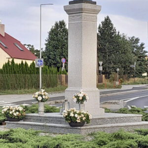 na wysokim kamiennym postumencie, ustawionym na prostokątnej kamiennej podstawie figura Matki Boskiej w biało-niebieskiej szacie z rozchylonymi dłońmi, za figurą fragment ulicy i dom mieszkalny