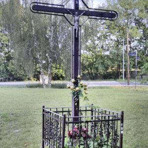 krzyż drewniano-metalowy z figurą ukrzyżowanego Chrystusa, otoczony metalowym niskim płotem stoi na trawniku, w głębi fragment ulicy i drzewa