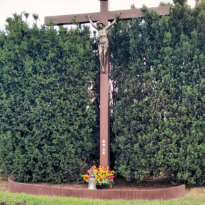drewniany krzyż z figurą ukrzyżowanego Chrystusa, nad nim tabliczka z napisem INRI, krzyż stoi w otoczeniu wysokich krzewów