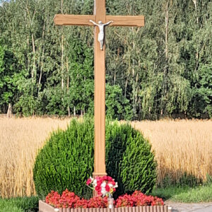 drewniany krzyż z metalową figurą ukrzyżowanego Chrystusa otoczony, przy nim rosną czerwone kwiaty, za nim zielony krzak na tle polnego krajobrazu i drzew