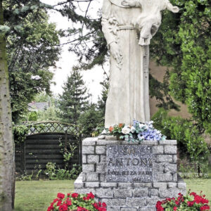 na ceglanym, pomalowanym postumencie figura świętego w habicie trzymającego na ręce dziecko, wokół postumentu kwiaty, w głębi drzewa i fragment drewnianego ogrodzenia