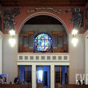 fragment wnętrza kościoła, nad wejściem balkon na dwóch filarach, na nim organy złożone z dwóch części - prawej i lewej, pośrodku nich okrągły kolorowy witraż, nad całością łuk, po obu stronach łuku malowidła