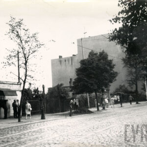 ogrodzona pusta przestrzeń między budynkami, chodnikiem spacerują ludzie, na pierwszym planie brukowana ulica