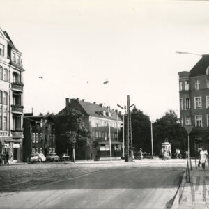 ulica, częściowo brukowana, przy ulicy kilkupiętrowe kamienice, spacerują ludzie