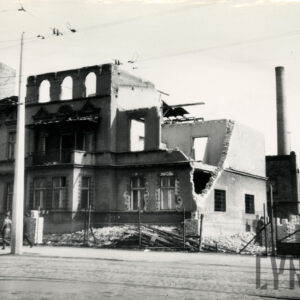 trzykondygnacyjny budynek w trakcie rozbiórki, wkoło budynku gruz i deski, budynek otoczony metalową siatką, za nim z prawej wysoki komin