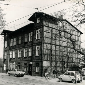 trzypiętrowy budynek o konstrukcji szkieletowej, drewnianej, wypełnionej cegłami, widok od ulicy, przy ulicy stoją dwa samochody osobowe, spacerują ludzie