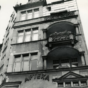 fragment elewacji kamienicy z dwoma z balkonami i wieżyczką na szczycie, na parterze kamienicy napis na ścianie „apteka”