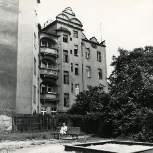 czterokondygnacyjna kamienica z poddaszem, przed budynkiem siedzi na ławce kobieta, przed nią piaskownica dla dzieci