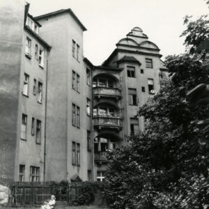 czterokondygnacyjna kamienica z poddaszem, przed nią na ławce siedzi kobieta