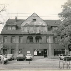 trzykondygnacyjny budynek z trójkątnym szczytem, przed nim zaparkowane samochody