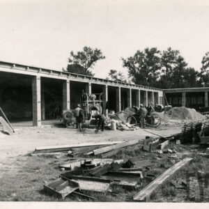 murowany, długi budynek z przedzielonymi boksami w trakcie budowy, przed teren budowy z betoniarką, kopce piachu i pracujący robotnicy