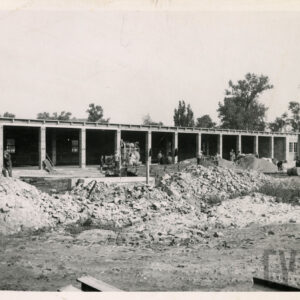 murowany, długi budynek z przedzielonymi boksami w stanie surowym, przed budynkiem gruz i pracujący robotnicy