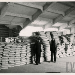 hala nakryta drewnianym skośnym dachem, w niej złożone jedne na drugim worki, obok nich stoi trzech mężczyzn