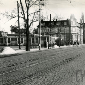 brukowana ulica z torami tramwajowymi, w głębi niski, przeszklony budynek z neonem na dachu, z nim kilkupiętrowe kamienice