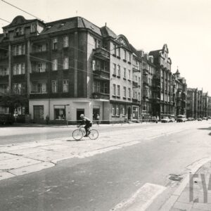 asfaltowa ulica z torami tramwajowymi, na narożniku trzypiętrowa kamienica z użytkowym poddaszem, w partii parteru sklepy, pośrodku ulicy jedzie rowerzysta