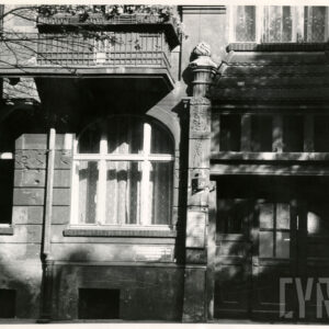 fragment elewacji kamienicy, na prawo drewnianych drzwi, po bokach dwa zdobione pilastry, wszystko zwieńczone daszkiem pokrytym dachówką, z lewej okno parteru, nad nim balustrada balkonowa pierwszego piętra