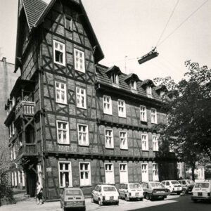 dwupiętrowy dom drewniano-ceglany z użytkowym poddaszem, narożnik z dodatkową kondygnacją, nakrytą stromym dwuspadowym dachem, przed budynkiem zaparkowane samochody