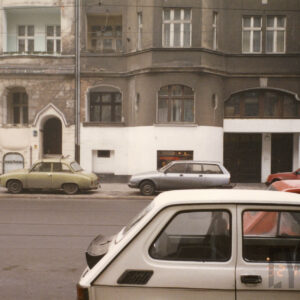 trzy pierwsze kondygnacje kamienic, na pierwszym planie ulica z zaparkowanymi na niej samochodami