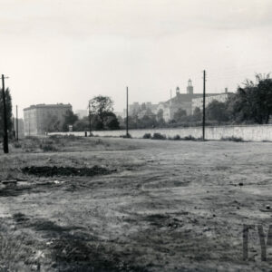 pusta przestrzeń, w głębi murowany płot, daleko kilkupiętrowe zabudowania, z prawej drewniany krzyż