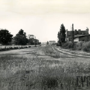 z lewej murowane ogrodzenie i fragment kamienicy, z prawej ceglany budynek, środkiem ciągną się nieużytki, na wprost w oddali kilkupiętrowa kamienica
