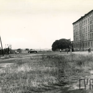 z przodu nieużytki, z prawej kilkupiętrowa kamienica narożna, spacerujący ludzie, na wprost stoi ciemny samochód