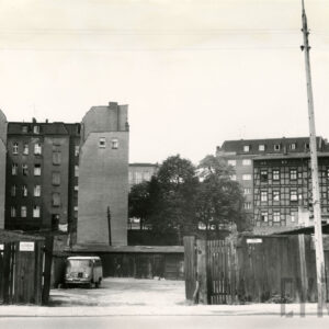 drewniane ogrodzenie z otwartą bramą na podwórze z drewnianymi szopkami, w głębi z lewej wielokondygnacyjne kamienice, z prawej dom drewniano-ceglany