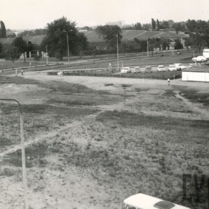 na pierwszym planie pusta przestrzeń, w głębi trybuny stadionu, z prawej stoją samochody i autobusy