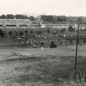 na pierwszym planie murowane baraki, w głębi wieża do skoków do wody i trybuna dla publiczności