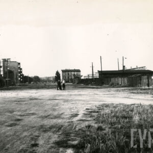 z przodu pusta przestrzeń, pośrodku dwóch stoi mężczyzn, z lewej bloki mieszkalne, na wprost kilkupiętrowa kamienica