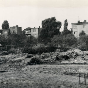 na pierwszym planie nieużytki, w głębi za krzakami i niskimi drzewkami jednorodzinne, murowane domy mieszkalne