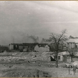 na pierwszym planie pusta przestrzeń ze składowiskami gruzu i śmieci, w głębi kamienice, na horyzoncie dym unoszący się z wysokiego komina