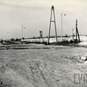 z przodu gruntowy teren, w głębi betonowy słup z przewodami elektrycznymi, za nim niski budynek z oknami