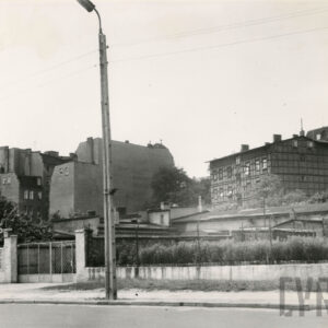 z przodu niska zabudowa, słabo widoczna za krzakami ogrodzona metalową siatką, w głębi kilkukondygnacyjne, otynkowane kamienice, z prawej dom drewniano-ceglany, tzw. mur pruski, w centrum na pierwszym planie wysoka lampa na betonowym słupie