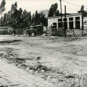 gruntowa droga, z nią murowany niski budynek w budowie, w głębi drzewa